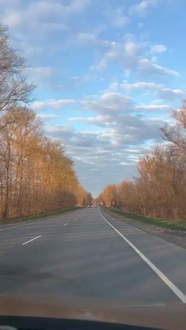 The video presents a first-person perspective from inside a vehicle driving on a straight, two-lane asphalt road. The road is bordered on both sides by trees with bare or sparse foliage, suggesting a season like late autumn or early spring. Above, the sky features a vibrant blue interspersed with scattered white cumulus clouds, arranged in horizontal bands. The lighting is warm and golden, indicative of either early morning or late afternoon, casting long shadows across the road.

Ahead in the distance, several vehicles are visible. A red truck is seen driving in the same direction as the viewer's vehicle. Later in the clip, a white Mercedes-Benz truck, equipped with a crane on its flatbed, passes by in the oncoming lane. A red triangular warning sign with a black exclamation mark is visible on the right side of the road. The scene conveys a serene drive through a rural landscape, with the dynamic sky as a prominent feature. The location context provided is Shiryaevo, Russia.