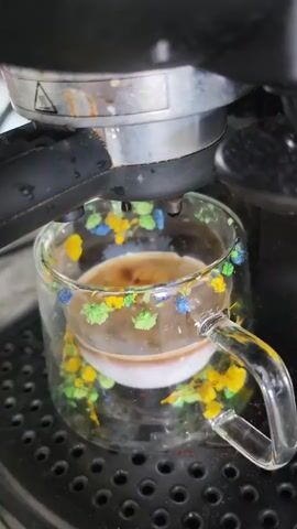 This is a close-up video of an espresso machine brewing coffee into a decorative glass mug. The mug is transparent and appears to have a design of colorful flowers or abstract shapes embedded within the glass walls. Two streams of dark espresso are pouring from the espresso machine's portafilter into the mug, creating a crema on top of a white liquid, likely milk. The espresso machine is black and metallic, with visible water droplets on its surface. The mug is placed on the drip tray of the espresso machine, which has a patterned surface. The lighting suggests an indoor setting. The video is a short loop, showing the coffee continuously brewing.