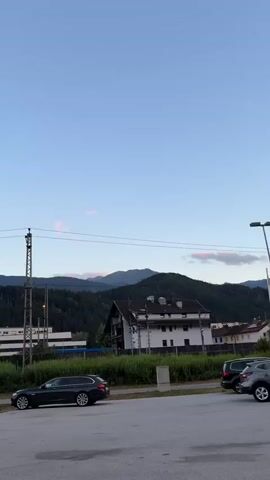 The video shows a landscape view at dusk. A dark-colored station wagon is parked in a paved lot. In the background, there are trees, buildings, and mountains under a clear sky. There are power lines and utility poles visible.