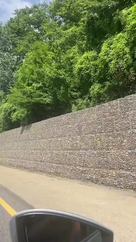 The video shows a car driving alongside a steep embankment reinforced with a gabion wall. The wall is constructed from wire baskets filled with rocks and is several tiers high, creating a stable retaining structure. Above the gabion wall, lush green trees and foliage cover the hillside, indicating a natural, possibly wooded, environment. The road surface is visible in the foreground, with a yellow line marking the lane. The sky appears partly cloudy. The perspective suggests the video is filmed from the driver's side of the vehicle, with a portion of the side mirror visible at the bottom of the frame. The overall scene depicts a man-made structure integrated into a natural landscape, likely a highway or road cut.