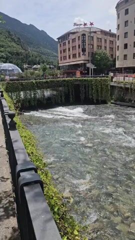 The video presents a stationary eye-level view of a clear river flowing from background to foreground under a bright, partly cloudy sky. In the immediate foreground on the left, a black metal railing borders a concrete walkway, with green vines growing along its base and posts. The river occupies the center and right, revealing rocks on its bed and showing visible current with small white rapids. On the left bank, a retaining wall covered in dense green vines extends across the frame, featuring a small waterfall-like cascade where the river water drops slightly. In the midground, beyond the river, a multi-story building, possibly a hotel, stands prominently with a red sign and two stars on its roof. Another building is partially visible to its right. Large, dark green mountains rise in the distant background. The scene is brightly illuminated by sunlight.