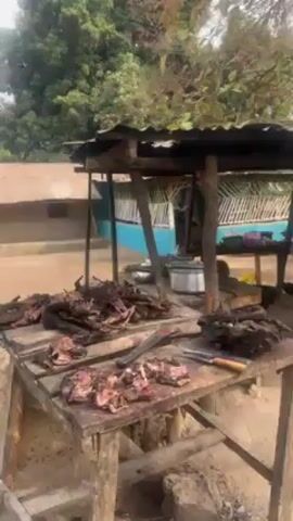 The video depicts an outdoor meat processing area in a rural setting. A weathered wooden table is prominently featured, covered with raw meat, some of which appears to be charred or smoked. A large cleaver and knife lie among the meat. Another table is partially visible in the background, along with large cooking pots and a rudimentary cooking fire. A building with blue trim is visible in the background, as well as a parked motorbike. The area appears to be sandy or dusty, and trees surround the scene.