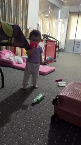 A young child stands in a room, holding a pink object to their mouth. The child is wearing a purple polka dot shirt and grey pants. A green bottle is on the floor nearby. In the background, there is a pink baby bed with a white blanket, a red playpen, and a pink suitcase. Curtains hang on a window to the left. The floor is carpeted.