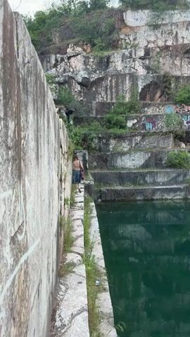 The video opens with a wide shot of a former quarry featuring tiered rock faces and a deep green body of water. A person, shirtless and wearing shorts, walks away from the camera along a narrow concrete path adjacent to the water, which is on the right. Graffiti marks the concrete wall on the left. In the background, the quarry walls rise in distinct, flattened rock tiers, interspersed with sparse vegetation and visible graffiti in various colors. As the video progresses, it zooms in on one of the mid-level rock tiers. On this tier, three camping tents are pitched, two light-colored and one darker. In the center of this tier, a small group of approximately three to four individuals is seated around a light-colored table, seemingly interacting and relaxing. The surrounding rock faces continue to display extensive graffiti.