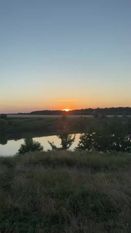 The video shows a sunset over a rural landscape. The sun is a bright orange orb near the horizon, casting a warm glow across the sky. The sky is a gradient of soft orange and yellow, with a few wispy clouds. Below the sun, a dark line of trees forms a silhouette against the bright sky. In the foreground, a body of water reflects the colors of the sunset. The water is calm, with subtle ripples. The banks of the water are covered in greenery, including trees and tall grass. The overall impression is one of peacefulness and natural beauty.