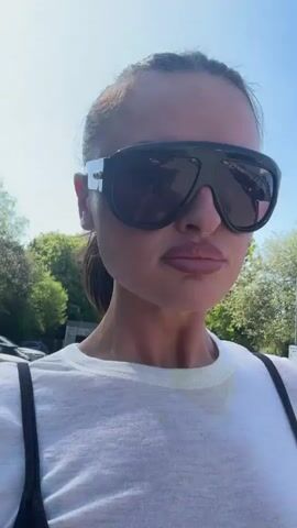 A woman is walking, and recording herself with a cell phone camera. She's wearing large sunglasses, a white t-shirt over a black tank top, and white pants. She has her hair pulled back. The background shows trees and a building. She is smiling and appears to be in a parking lot. The sunlight is bright.