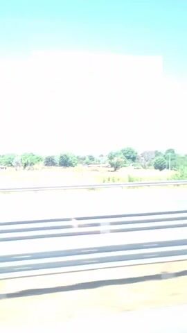 The video shows a roadside view from a vehicle in motion. The scene is a sunny day with a clear blue sky. In the foreground, there are highway guardrails and a blurred green landscape indicating movement. Beyond the highway, there is a rural landscape with trees, vegetation, and some buildings in the distance. A tall electrical pylon is visible on the right side of the frame at the beginning of the video and moves to the left as the perspective shifts. The reflection of the sky and possibly clouds or internal vehicle elements is visible on the window, creating abstract shapes. There are no people visible, and the action is limited to the passive observation of the passing scenery.