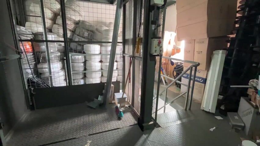 The camera is positioned inside a service elevator or lift. The lift is loaded with stacks of wrapped cylindrical items, likely industrial materials, filling the space behind a wired cage.  To the right of the cage, outside the lift, is a landing area. It is cluttered with cardboard boxes, a ladder, and other assorted items. The floor of the lift is diamond-patterned metal. The camera then abruptly tilts upwards.