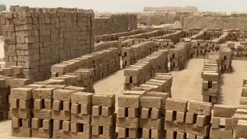 The video depicts a vast brickyard filled with stacks of freshly made bricks. The bricks are arranged in rows and columns, covering a large expanse of sandy ground. The stacks of bricks are primarily brown, with slight variations in color. The landscape appears arid and dusty, under a hazy sky. There are no people visible in the frame.
