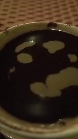 The video shows a close-up, top-down view of a dark liquid in a light-colored cup. The liquid appears to have a foamy, lighter-colored substance floating on top, creating irregular shapes. The background is a textured, brown surface, possibly a placemat or a woven material. The lighting is dim. The cup itself has a textured, possibly ceramic exterior. There are no people or discernible actions taking place. The focus is solely on the contents of the cup.