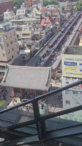 This is a high-angle, vertical video of a busy street market in a city. The market is lined with stalls under awnings, with many people browsing. In the foreground, a black metal railing is visible, suggesting the viewpoint is from an elevated position like a balcony or rooftop. The buildings surrounding the market appear to be a mix of traditional and modern architecture, with some taller buildings in the distance. A large advertisement billboard with a cartoon character is also visible on the side of a building. The sky is blue with some clouds. The video pans slightly upward throughout its duration, revealing more of the cityscape.