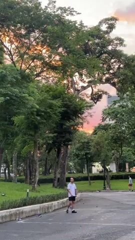 The video shows a park with large trees and green grass. The sky has pink and orange hues, indicating sunset. In the foreground, a man in a white t-shirt and black shorts stands on a paved surface, looking towards the camera. There are some objects, possibly shoes, scattered on the ground near him. In the background, another person is walking away from the camera, and a building is visible behind the trees. The scene appears to be a tranquil outdoor setting at dusk.