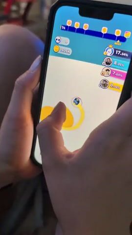 The video shows a close-up of two hands holding a smartphone. The phone screen displays a colorful mobile game with graphics that appear to be animated. The game involves a yellow blob-like object and a small circular avatar, with progress bars and player rankings visible at the top of the screen. One hand's thumb is positioned on the lower left side of the screen, seemingly interacting with a smaller blue and pink object. The other hand's fingers are curled around the right side of the phone, with the index finger also appearing to touch the screen near the yellow blob. The background is blurred, but suggests an indoor setting with some reddish furniture visible. The lighting is dim, with the phone screen being the primary light source.