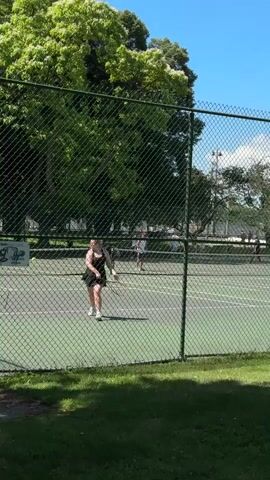 A video shows a tennis court with several people playing tennis. A woman in a black dress is in the foreground, hitting a tennis ball. A man in a red shirt is in the background, also playing tennis. The court is green, and there is a fence around it. There are trees and a blue sky in the background. The video is filmed from behind the fence.