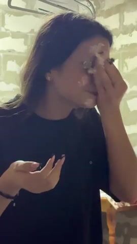 A young woman with long brown hair is in the process of applying something white and creamy to her face. She is wearing a black t-shirt. Her left hand is holding a piece of cake or similar item, while her right hand is smeared with the white substance as she applies it near her eye. She is looking to her right, possibly at someone filming her. In the foreground, another hand is holding up a smartphone, which appears to be recording her. The background shows a brick wall and a window with curtains.