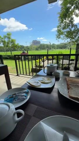 Here is a description of the video:
The video shows a breakfast spread on a table on a covered patio. The table has plates of eggs, toast, and meats along with tea and condiments. Beyond the table is a black metal fence, then a grassy field. In the distance is a large rock formation. The sky is blue with white clouds. There are trees on the right side of the video.
