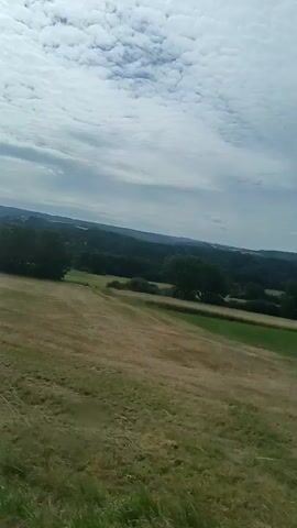 This is a first-person perspective video from inside a vehicle, likely a van or truck, as it drives through a rural landscape. The camera is positioned to look out the windshield and side window. The scene outside consists of rolling hills covered in fields of dry grass or hay, interspersed with patches of trees and a winding road. The sky is cloudy with patches of blue. The interior of the vehicle is visible in the foreground, including the dashboard, a sun visor, and a side mirror. Some cans are visible in a compartment on the dashboard. The vehicle appears to be in motion on a paved road.