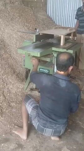 Two men are in a woodworking shop. One man, wearing a dark shirt and plaid shorts, is crouched down in front of a green woodworking machine and appears to be operating it. The other man, wearing a striped shirt and beige pants, stands behind the machine. The shop is filled with wood shavings, piled high against the back wall and on the floor. The machine has a flat surface with various attachments and a wooden jig. The lighting suggests it is daytime.