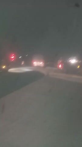 This media file depicts a blurry nighttime scene on what appears to be a road or airport tarmac in Sabon Gari, Nassarawa, Nigeria. The setting is outdoors, with a dark, overcast sky. Several vehicles are visible, including what looks like a white truck with red taillights in the foreground to the left, and other vehicles further in the distance, all illuminated by their headlights and taillights, creating streaks of light due to the blur. There are also some streetlights or other fixed lights in the background. The ground appears to be a light-colored, possibly paved surface, with some darker areas suggesting shadows or different materials. The overall impression is one of movement or transit at night. There are no discernible people in the frame. The atmosphere is dim and somewhat indistinct due to the low light conditions and the motion blur. There is no visible text in the media file.
