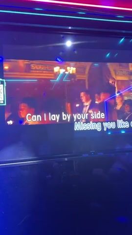 A karaoke room featuring a large screen displaying lyrics is shown. The room has blue neon lighting. A person is sitting on a couch, looking at their phone. Another person is standing and singing into a microphone. The singer is wearing a black top and light-colored jeans. There are drinks and accessories on the table. The room is dimly lit, with a strong focus on the screen and neon lights.
