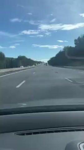 The video depicts a car driving on a highway, visible from the driver's perspective. The highway has multiple lanes and is marked with white lines. Other vehicles, including cars and trucks, are visible in the distance. The sky is blue with scattered clouds. Trees line both sides of the highway. The dashboard of the car is visible in the foreground. Someone is speaking, presumably inside the car, but the content is not discernible.