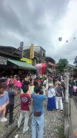 The video shows a crowded street in what appears to be a town or city. People are standing on and around train tracks, many holding up their phones to take photos or videos. There are buildings with shops and restaurants lining the street, some with signs in Asian languages. In the sky, many sky lanterns are floating upwards. The sky is overcast. People are interacting with each other, pointing at the lanterns, and generally observing the scene.