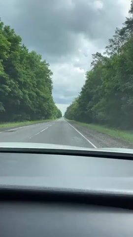 The video is captured from inside a vehicle, looking forward through the windshield. A two-lane asphalt road stretches ahead, flanked by dense green trees on both sides. The sky is overcast with dramatic, dark clouds. The perspective suggests the vehicle is in motion, traveling down the road. The dashboard of the vehicle is visible in the foreground.