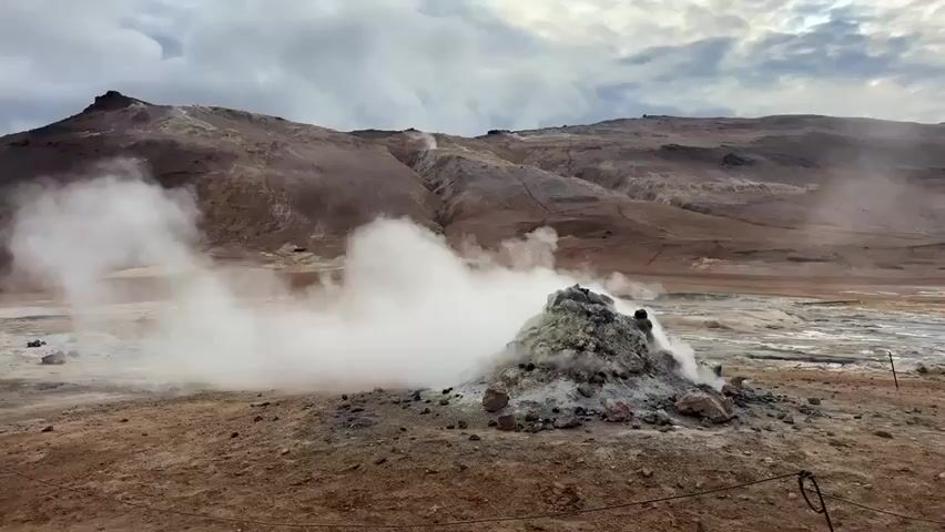 The video captures a wide shot of a geothermal field in Reykjahlíð, Iceland, characterized by active steam vents and barren terrain. In the foreground, a prominent rocky mound, stained with various mineral colors, vigorously emits a large plume of white steam that drifts to the left. Further into the midground, several smaller plumes of steam rise from other vents and mud pots, indicating widespread geothermal activity across the landscape. The ground is a mix of reddish-brown earth, grey rock, and lighter patches, possibly of sulfur or other mineral deposits.

The background features rugged, barren mountains with a brownish hue, extending across the horizon under an overcast sky filled with grey clouds. No people are visible in the scene. A thin wire or rope fence is subtly visible in the bottom right corner, suggesting a designated viewing area for visitors. The overall impression is one of raw, powerful geological forces at play in a stark and otherworldly environment. There is no visible text.