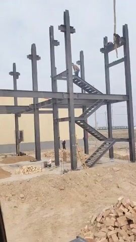 The video shows a construction site from the interior of a vehicle, possibly a crane or other heavy machinery. The main structure being built appears to be a steel frame building, with vertical columns and horizontal beams in place. Workers are visible on the steel structure and at ground level. There are piles of bricks and dirt on the ground. The vehicle has screens and controls visible, suggesting it's an operating machine. The background reveals other industrial buildings and a flat, open landscape.