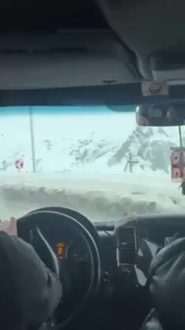The video shows a view from the interior of a car driving through a snow-covered landscape. The dashboard, steering wheel, and driver’s hands are visible. A Georgian flag ornament hangs from the rearview mirror. Outside the windshield, snow-covered mountains, some buildings, and a road sign are seen. The sky is overcast. Another vehicle passes on the road.
