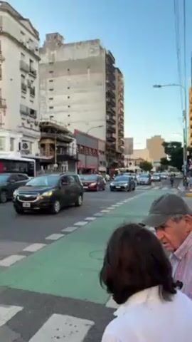 The video shows a street scene with traffic in Buenos Aires, Argentina. The scene is filmed from the perspective of someone standing on the corner of a crosswalk. In the foreground, a man and a woman stand waiting to cross the street. The man is wearing a cap and a red-and-white plaid shirt. The woman is wearing a white shirt. Traffic is moderate, and includes cars, motorcycles, and a bicyclist. The buildings along the street are tall and typical of urban architecture. The crosswalk is marked with a green painted lane. The sky is blue and clear, suggesting either late afternoon or early morning.