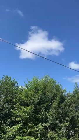 The video shows a view of a bright blue sky with scattered white clouds. A thin black wire runs diagonally across the frame. In the lower part of the video, there are green trees and foliage. A narrow road is visible in the distance, with a small building and a white car parked nearby. The scene appears to be outdoors on a sunny day.