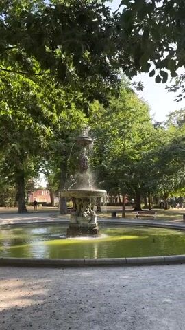 The video displays a fountain in a park. The fountain is ornate and features multiple tiers with water cascading down. The water in the basin has a greenish hue. Lush green trees with dense foliage surround the fountain, creating a shaded environment. Sunlight filters through the leaves, casting dappled shadows on the ground. In the background, there are hints of buildings and park benches. A single person is visible walking in the distance. The overall atmosphere appears serene and natural.