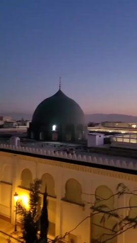 The video shows a view from a height, looking out over a building complex with a large green dome, likely a mosque, set against a twilight sky. The sky transitions from a deep blue overhead to a blend of orange and pink near the horizon, where the silhouettes of distant mountains are visible. Below, the architecture features arches and white-colored structures, illuminated by yellow lights. Scattered lights suggest urban development in the distance. A partially obscured tree is visible in the foreground. The camera pans slowly to the right, revealing more of the building complex and cityscape.
