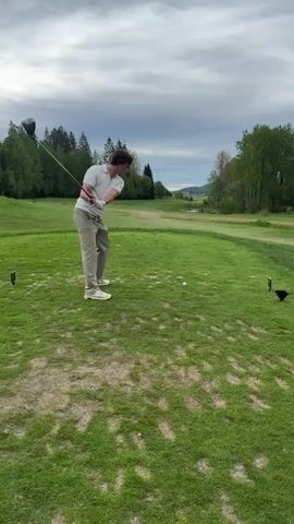 A man in a white t-shirt and khaki pants hits a golf ball with a driver on a grassy tee box. The man is right-handed. After hitting the ball, he watches its trajectory. The sky is overcast. The tee box is surrounded by green grass, with trees in the background. There is a glimpse of water in the distance.