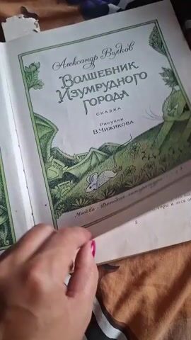 The video shows an open book with illustrations. On the left page, an illustration depicts a scene with witches and bats over a cauldron of bubbling liquid. The style is dark and fantastical. The right page contains text with a heading "Ураган" (Hurricane). The illustration above the text shows a group of figures in a chaotic scene. The book is lying on a surface that appears to be a patterned blanket or bedding. A person's hands with pink nail polish are visible turning the pages. The lighting suggests it might be daytime, but it's difficult to determine the exact time of day from the image. The setting appears to be indoors, possibly a home. The text is in Russian and describes a hurricane. Some of the visible text mentions "фермер Джон" (farmer John) and "девочка Элли" (girl Ellie). The overall mood evoked by the illustrations and the title of the text is one of suspense and possibly danger.