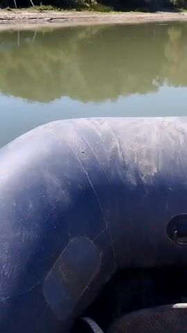 The video begins with a close-up of a deflated, dark gray inflatable boat. The camera then pans out to reveal a body of water, likely a lake or river, with a shoreline in the background. The shoreline is covered in lush green trees and vegetation. On the shore, there is a blue tarp supported by poles, possibly serving as a makeshift shelter, and some scattered debris. The water is calm and reflects the trees and sky. The overall scene suggests a natural outdoor environment, possibly a campsite or a recreational area.