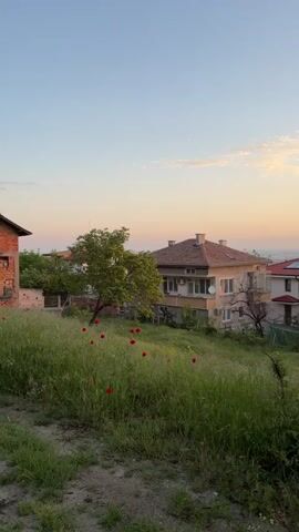 The camera pans slowly across a residential landscape in the early morning or late evening, given the soft, warm hues of the sky. On the left is an unfinished brick building with open window spaces, next to a tree. To the right is a completed two-story house with a tiled roof and a balcony. Tall green grass and weeds cover the hillside between the viewer and the houses, interspersed with vibrant red poppies. An unpaved dirt path is visible in the foreground. Power lines are stretched across the top of the frame against the pastel sky. In the distance, more houses can be seen, suggesting a suburban or rural setting. The overall atmosphere is peaceful and quiet, with a hint of rustic charm.