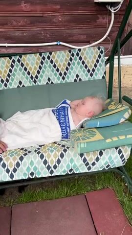 A person is sitting on a swing, and a baby is sleeping next to them. The swing is outside, and there is grass and a wooden building in the background. The person is wearing a light blue t-shirt and gray sweatpants. The baby is wrapped in a white blanket and is wearing a blue and white towel. The swing has a colorful patterned cushion. The person reaches out and touches the baby's blanket.