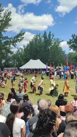 The video shows a crowded outdoor event, possibly a festival or gathering, with many people watching a performance. In the foreground, a diverse crowd of spectators, mostly adults, are seated or standing, facing away from the camera. Their backs are to the viewer, and their faces are mostly obscured. They are dressed in casual summer clothing. In the middle ground, a group of individuals, some in traditional Indigenous regalia, are dancing in a circular formation on a grassy field. They are adorned with colorful clothing, feathered headdresses, and beaded accessories. Some are holding objects, possibly drums or rattles. Behind the dancers, a large white tent is set up, and numerous flags are displayed, suggesting a ceremonial or organized event. The background consists of lush green trees and a bright blue sky with scattered white clouds, indicating a sunny day. The overall atmosphere appears festive and celebratory.