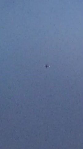 The video shows a drone flying in a clear, overcast sky. The drone is small and appears to be in the center of the frame. It has lights on its underside, which are illuminated. The drone is not moving relative to the camera, suggesting it is either hovering or the camera is tracking it. The background is a uniform blue-gray color. There are no other objects or people visible in the video. The footage is grainy and appears to be captured with a handheld camera or a camera with limited zoom.