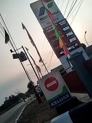 A video of a Pertamina gas station on a slightly overcast day. The camera is mounted on a moving vehicle, indicating traffic on a road. A tall Pertamina sign displays fuel prices, including Pertamax Turbo at 21650 and Dexlite at 20150. A red "stop" sign with the word "KELUAR" (exit) in green is visible in the foreground, implying the vehicle is near an exit. Several motorcycles are seen on the road, and a truck is driving away from the camera. The sky is a pale orange, suggesting it may be sunrise or sunset.