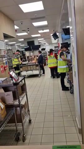 The video shows the interior of a McDonald's restaurant, likely in the back-of-house area. Several employees wearing yellow safety vests are packing food orders. One employee is standing by a cart with a brown paper bag and is looking at what appears to be an order sheet. Another employee is standing near a metal cart with a flat surface and is packing items into brown boxes. A man in a suit is standing near a cart filled with items and looking towards the back of the area. There are several TV screens mounted on the wall in the back. A metal rack with boxes and equipment is visible in the foreground on the left. The floor is tiled in a light color.