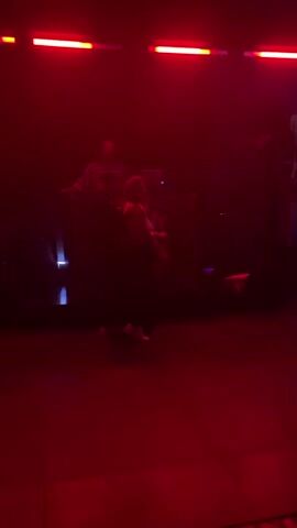 A man and a woman are slow dancing in front of a DJ booth in a club. The DJ is visible behind the booth. The lights are red and green. The woman is wearing a light colored top and the man is wearing a black suit. The dance floor is dark.