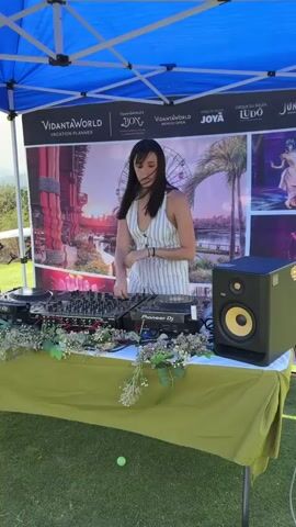 The video shows a DJ playing music under a blue tent on a golf course. A woman with long, dark hair is operating the DJ equipment, which includes two Pioneer DJ turntables and a mixer. The table is decorated with greenery. Behind her is a promotional banner for VidantaWorld. The setting is a golf course with a scenic view. A couple stands nearby under the tent, watching and chatting. Other people are seen in the background in golf carts. The sky is blue with some clouds.