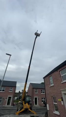 A telescopic handler with its boom extended vertically high into the air is positioned in a construction site between two identical brick houses. The handler is yellow and black, and its forks are visible at the top of the extended boom. The sky is overcast with clouds, and a lamppost is visible on the left side of the frame. The ground around the handler is rough and appears to be unpaved. There are no people visible in the video.