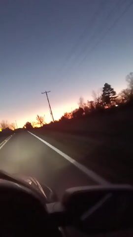 The video shows the view from inside a car driving on a road at sunset. The sky is a mix of blue and orange. There are trees and telephone poles along the side of the road. The car's headlights are on. The road has a white line marking the edge.