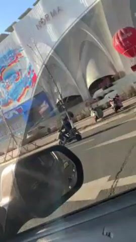 The video is filmed from the inside of a car driving on a road. The camera is focused on the street outside the passenger side window. The view includes the car's side mirror and the reflection of the car's interior in the window. 

A large building is visible in the background with a mural painted on its side. The mural consists of bold swirls and splashes of blue, red, and white paint. Above the mural, the building's name is written in Chinese characters. Solar panels are visible on the building's roof.

Other cars and a person riding a small motorbike pass by on the road outside. The weather appears to be sunny and clear.