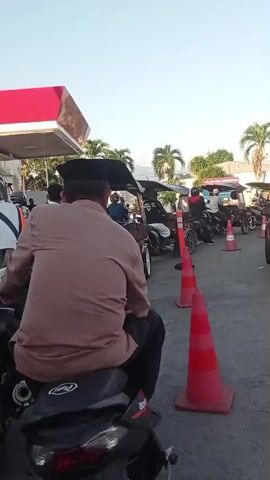 The video captures a busy scene at a gas station (SPBU), likely in Gorontalo, Indonesia, given the "DM" prefix on the license plate. Vehicles, predominantly motorcycles and motorized tricycles (with sidecars), are queued up, suggesting they are waiting to refuel. Red traffic cones are used to organize the line.

In the foreground, a man wearing a brown shirt and a black cap is seen from behind, sitting on a black motorcycle, also waiting in the queue. To his right, a red Honda Brio RS hatchback (license plate DM 1079 EC) drives past the queue and heads towards the pumps. Further along, a blue van-like vehicle is parked under the gas station's red and white canopy, possibly being fueled.

The setting features palm trees and other foliage in the background, under a clear, bright sky, indicating good weather. A gas pump island with the number "3" is visible under the canopy. Parts of a gas station sign with text like "DELAPAN... MANDIRI" can be faintly seen, typical of Pertamina stations. The overall atmosphere appears orderly, with people patiently waiting their turn.