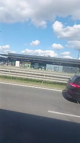 This video captures a roadside view of a commercial strip under a bright, cloudy sky, suggesting it's daytime with partly cloudy weather. The scene depicts a row of storefronts with large glass windows, typical of a modern retail complex. The stores visible include "STARBUCKS," "Vision Express," "RITUALS...", and "APTEKA" (which means pharmacy in Polish). Further down, "BLUMI" is also discernible.

Several vehicles are observed: a red car is prominent in front of the "APTEKA" store, a black SUV drives past the "RITUALS..." section, and a white van is parked near "APTEKA." In front of the "Vision Express" store, a person on a bicycle is visible, and other individuals can be seen through the windows, suggesting activity within the shops. The overall impression is one of a functional, commercial area, likely a shopping center or retail park.

The presence of Polish text ("APTEKA") and the provided location context (Mysiadło, Poland) place the scene specifically in Poland. The lighting indicates a sunny day, possibly mid-morning or afternoon, and the clouds suggest dynamic weather. The scene is captured from a moving vehicle on a road, indicated by the blurred asphalt and guardrail in the foreground. There are no strong emotional cues; the atmosphere is neutral and observational.